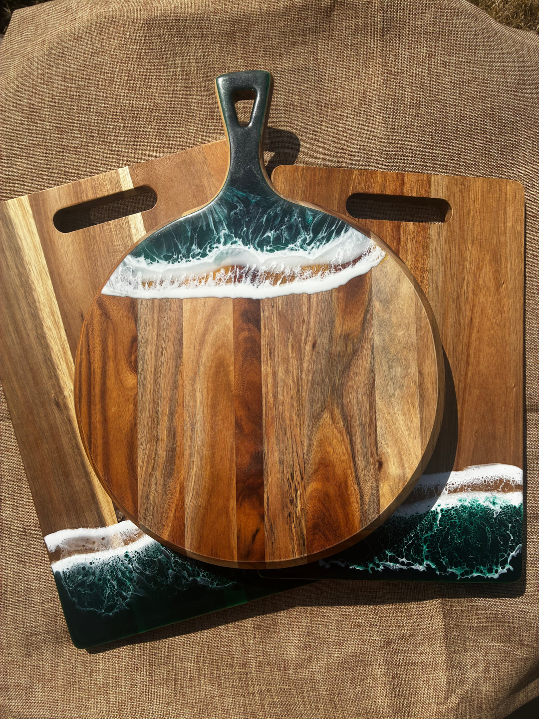 A round wooden serving board with a dark metal handle, featuring an ocean wave design in teal, white, and beige resin around the top and bottom edges, displayed on top of a rectangular wooden board with cutout handles.