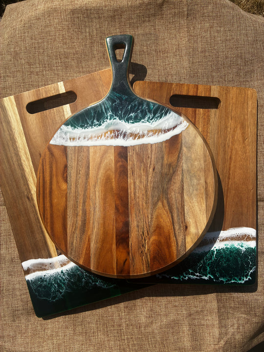 A round wooden serving board with a dark metal handle, featuring an ocean wave design in teal, white, and beige resin around the top and bottom edges, displayed on top of a rectangular wooden board with cutout handles.