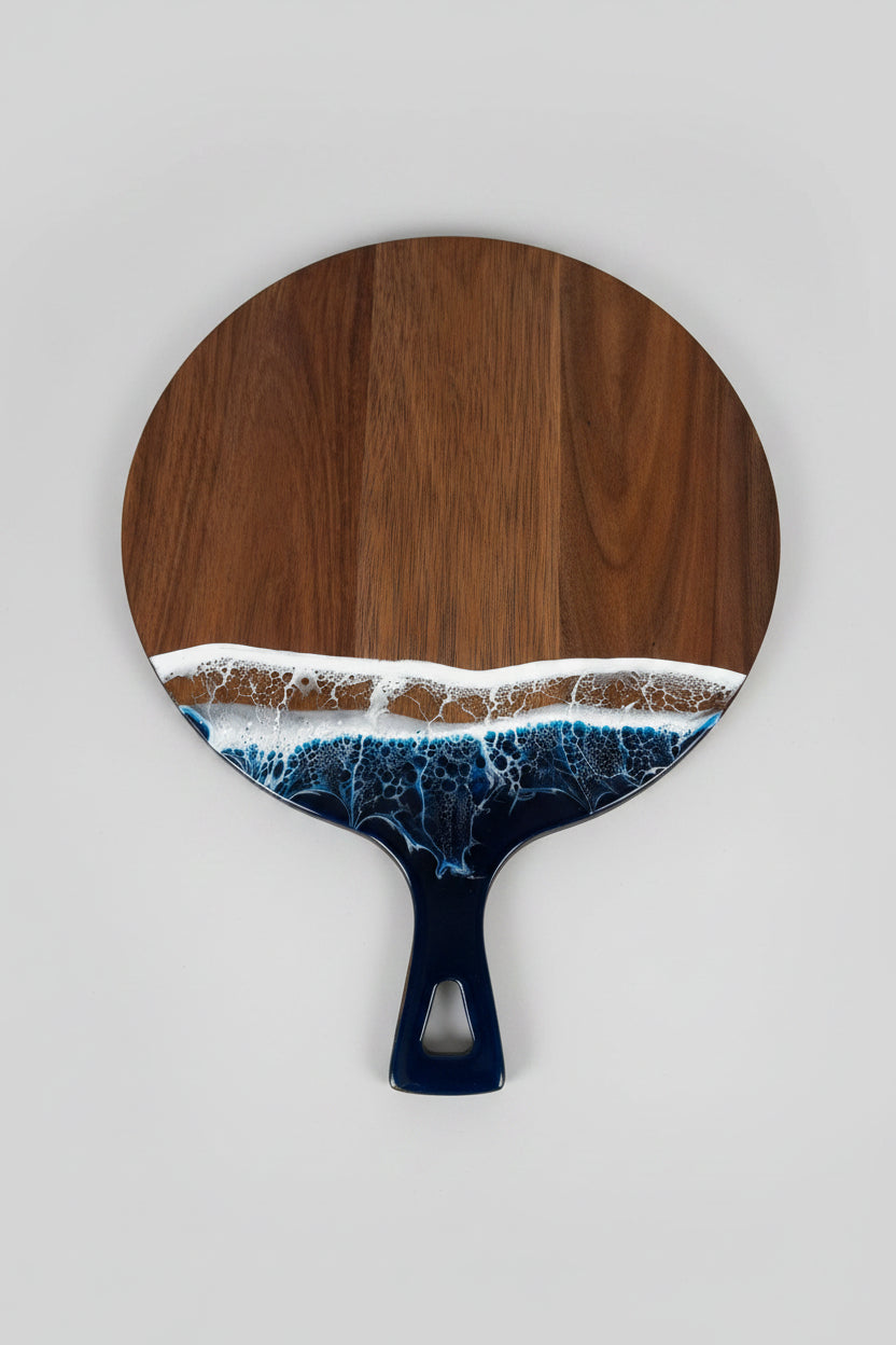 A paddle-shaped wooden board with a dark brown wood grain top and handle, featuring an embedded wave design in white and blue resin along the lower edge, resting on a textured white blanket.