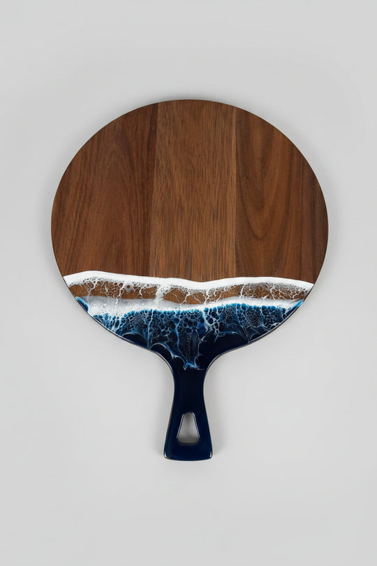 A paddle-shaped wooden board with a dark brown wood grain top and handle, featuring an embedded wave design in white and blue resin along the lower edge, resting on a textured white blanket.