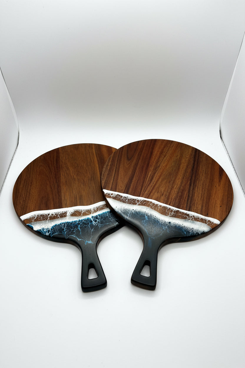 Two round wooden paddle-shaped items with black handles, featuring a wave design in blue, white, and brown resin along the bottom half, displayed on a textured cream fabric background.