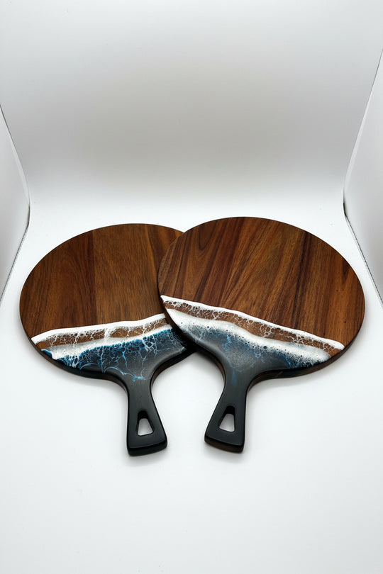 Two round wooden paddle-shaped items with black handles, featuring a wave design in blue, white, and brown resin along the bottom half, displayed on a textured cream fabric background.