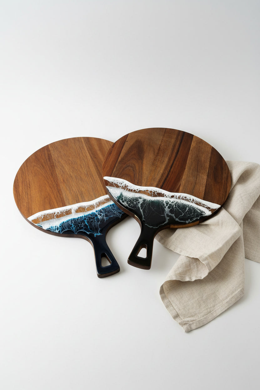 Two round wooden paddle-shaped serving boards with black handles, featuring a wave design in blue, white, and brown around the lower half, displayed on a textured white surface.