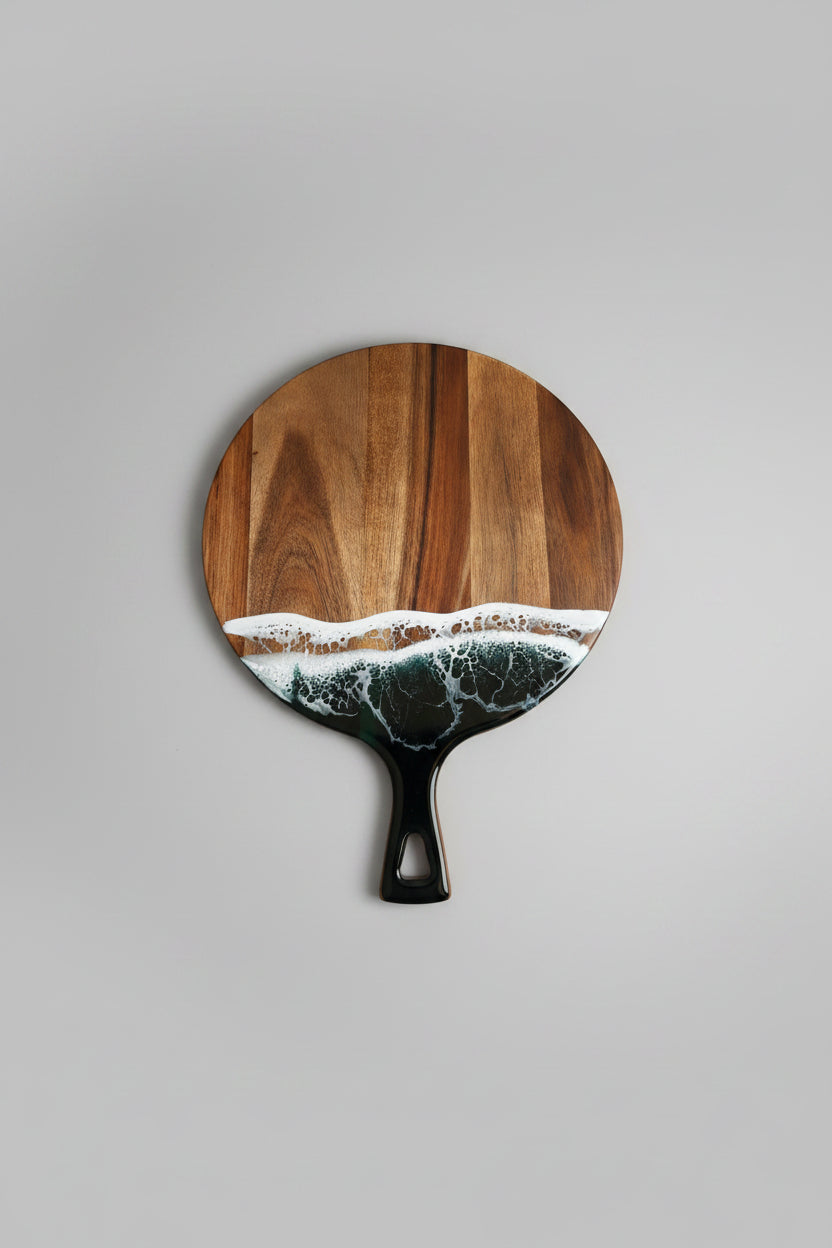 A round wooden serving board with a handle, featuring a printed or inlaid wave design in white, tan, and dark green near the bottom, set against a plain light gray background.