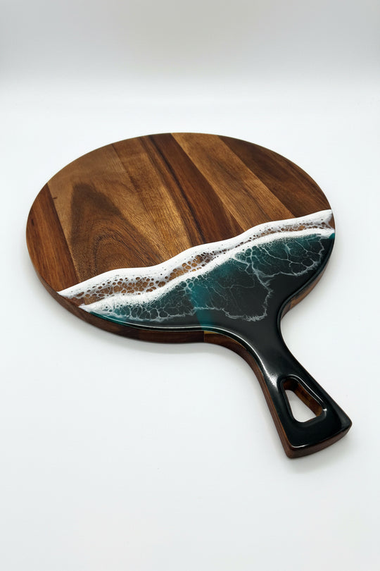 A round wooden serving board with a handle, featuring a wave design in the center. The wave has dark teal water, white foam, and a sandy beige shore, set against a polished dark brown wood grain background.
