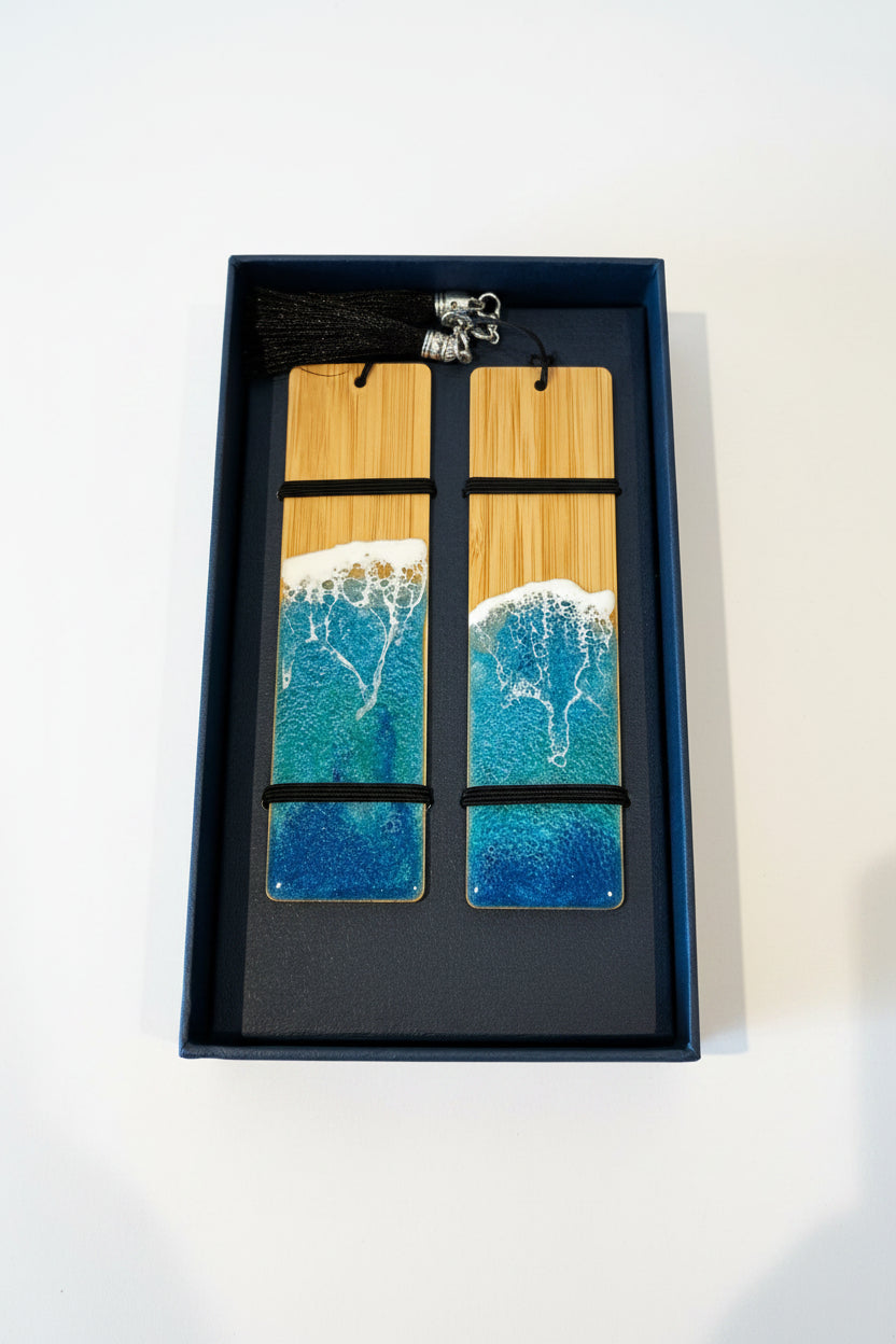 Two rectangular wooden bookmarks with a printed ocean wave design in blue, teal, and white, resting in a black presentation box with a black tassel.