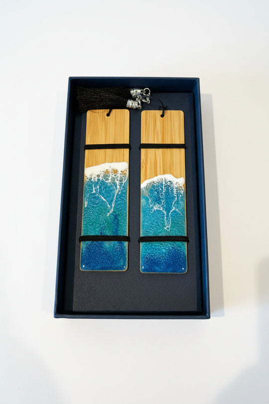 Two rectangular wooden bookmarks with a printed ocean wave design in blue, teal, and white, resting in a black presentation box with a black tassel.