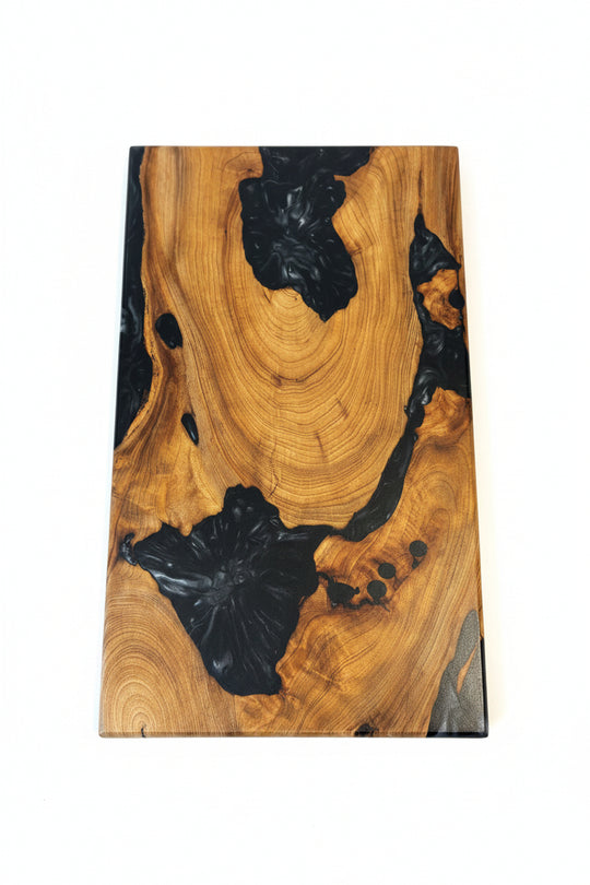 A rectangular live-edge wooden board with a rich golden-brown wood grain and glossy black resin pockets, displayed on a textured cream-colored towel.
