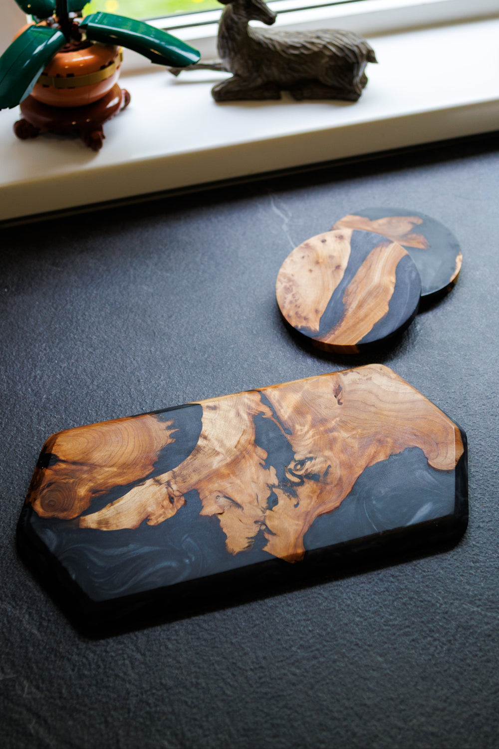 Two decorative coasters with a natural wood pattern on a dark surface, with a window and decorative items in the background.
