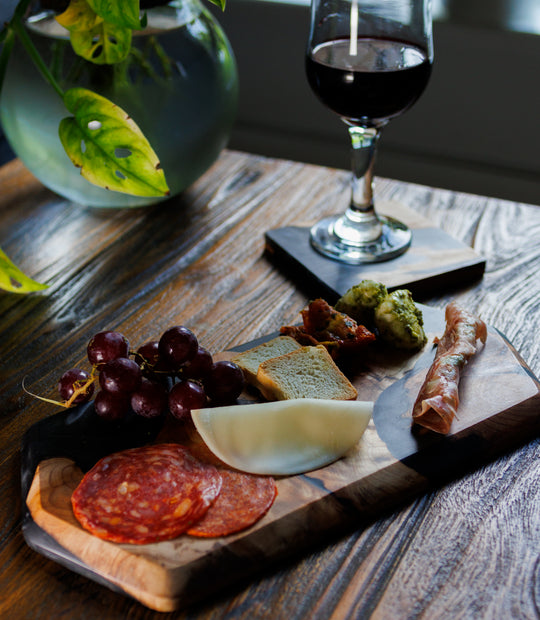 Wooden platter with cured meats, cheese, and grapes on a wooden table with a glass of red wine.
