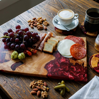 A styled charcuterie board on a rustic wooden table, featuring a wooden board with red marbled resin patterns, sliced salami, cheese, crackers, grapes, nuts, pickles, and a cup of coffee and tea nearby.