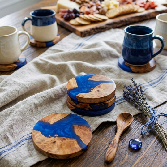 A styled tabletop scene with ceramic coasters and matching mugs in a home setting. The main sellable items appear to be the round wooden coasters with glossy blue resin inlay, shown in use on a wooden table beside a charcuterie board, cloth napkin, spoon, and dried lavender.