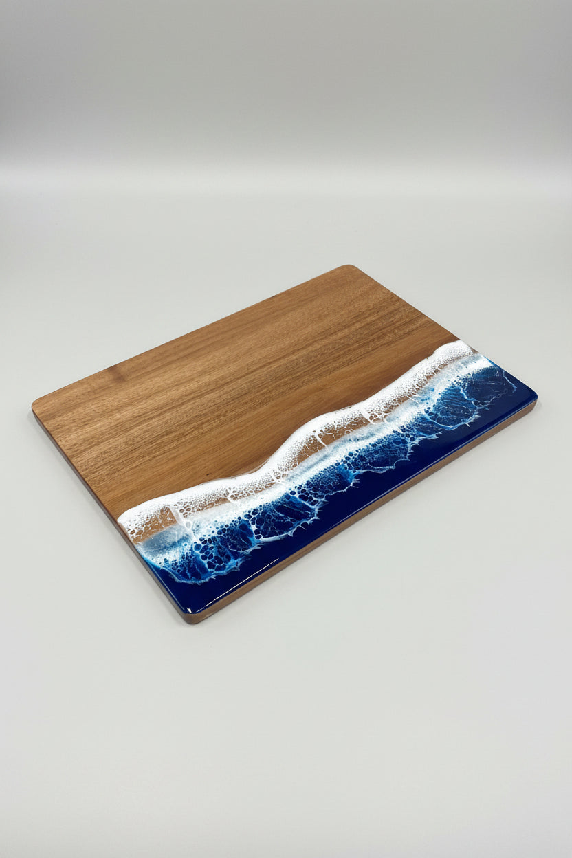 A rectangular wooden cutting board with a wave-like resin design in blue, white, and tan along the bottom half, placed on a wooden floor.