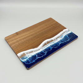 A rectangular wooden cutting board with a wave-like resin design in blue, white, and tan along the bottom half, placed on a wooden floor.
