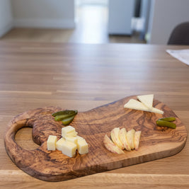A wooden charcuterie board with cheese wedges and slices, pickles, and two cocktails on a wooden table.