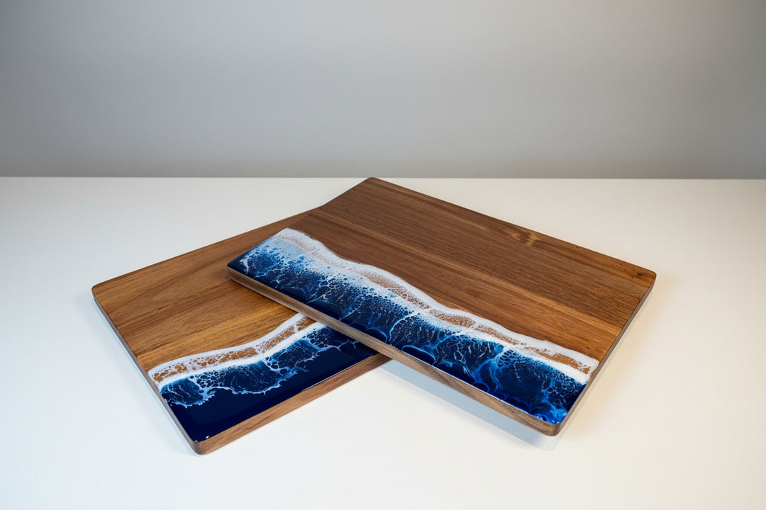 Two rectangular wooden serving boards with a blue and white ocean wave resin design on the bottom half, displayed on a wooden table with a green cocktail in the background.