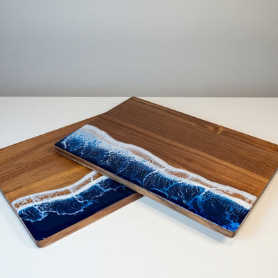 Two rectangular wooden serving boards with a blue and white ocean wave resin design on the bottom half, displayed on a wooden table with a green cocktail in the background.