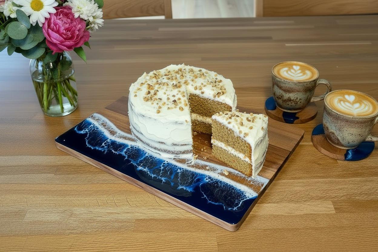 A rectangular wooden serving board with a blue and white ocean wave resin design on one end with a cake on it, placed on a wooden table next to two coffee cups and a vase of flowers.