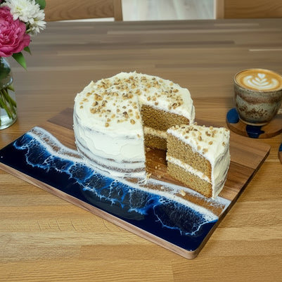 A rectangular wooden serving board with a blue and white ocean wave resin design on one end with a cake on it, placed on a wooden table next to two coffee cups and a vase of flowers.