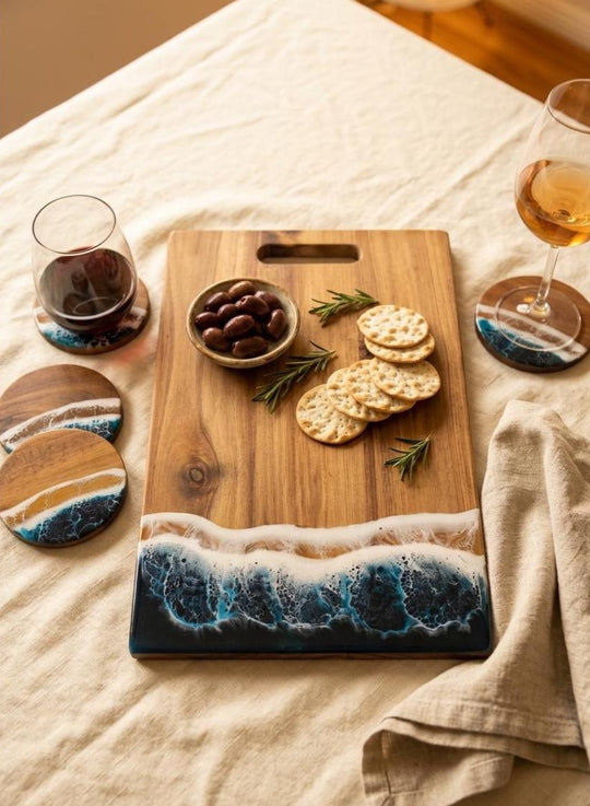 A rectangular wooden serving board with a wave design in blue, white, and dark navy resin at the bottom, styled on a beige linen tablecloth with crackers, olives, rosemary, and coasters around it.