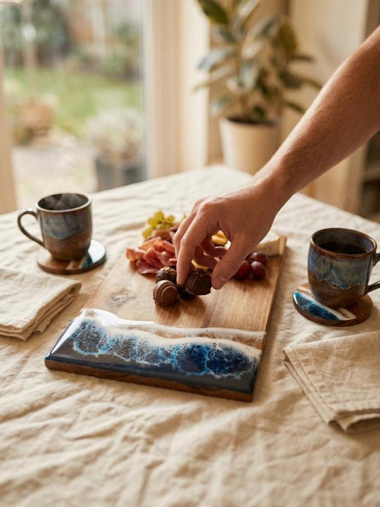 A ceramic serving board with a blue and white wave pattern on one end, placed on a beige tablecloth. A hand is placing nuts on the board, which also contains sliced fruit and crackers.