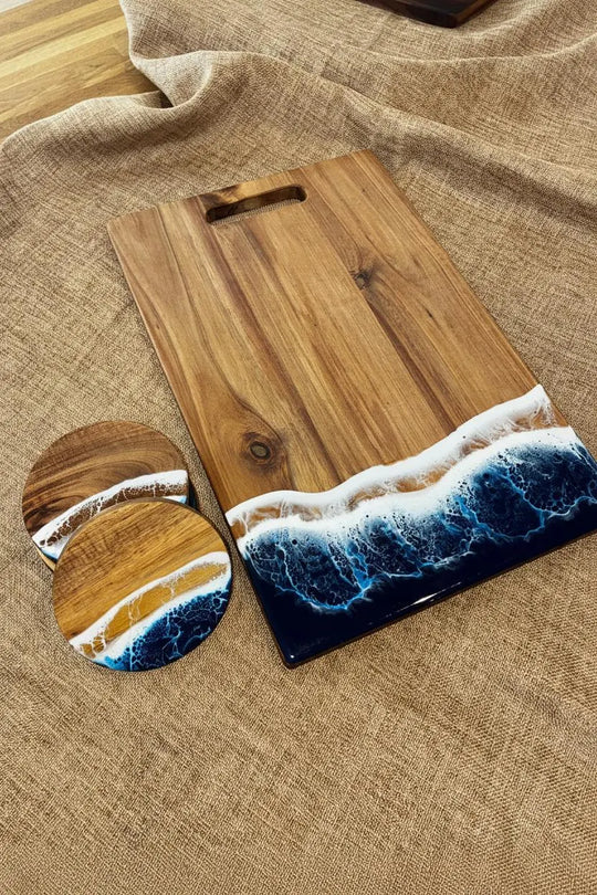 A rectangular wooden cutting board with a handle, shown on a beige linen surface alongside two round coasters. The board and coasters feature a printed or embedded ocean wave design in blue, white, and brown wood tones.