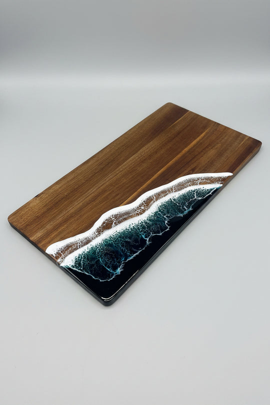 A rectangular wooden serving board with a glossy resin wave design in teal, white, and black, placed diagonally on a textured beige fabric surface.