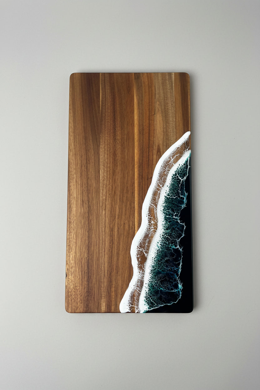 A rectangular wooden serving board with a dark brown wood grain finish, featuring an embedded wave design on the right side that mimics the shoreline with white foam, tan sand, and deep blue-green water.