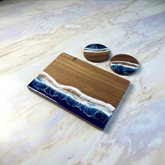 A rectangular wooden serving board with a blue and white ocean wave resin design along one end, shown on a light marble surface with two matching oval coasters behind it.