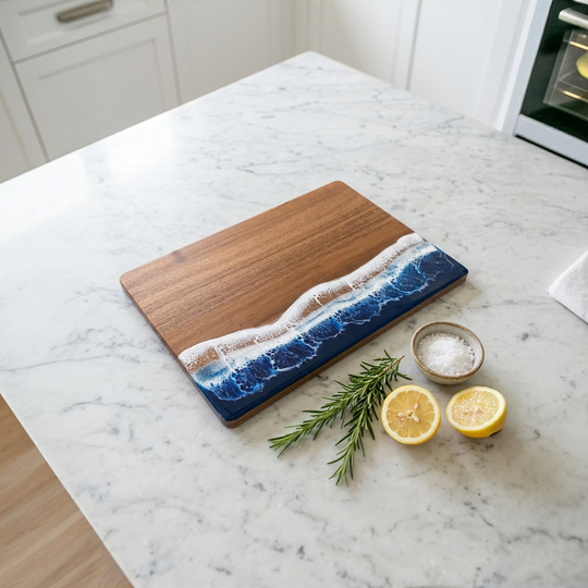 A rectangular wooden cutting board with a blue ocean wave and white foam design on one side, placed on a white marble kitchen countertop with lemon halves, a rosemary sprig, and small bowls of salt.